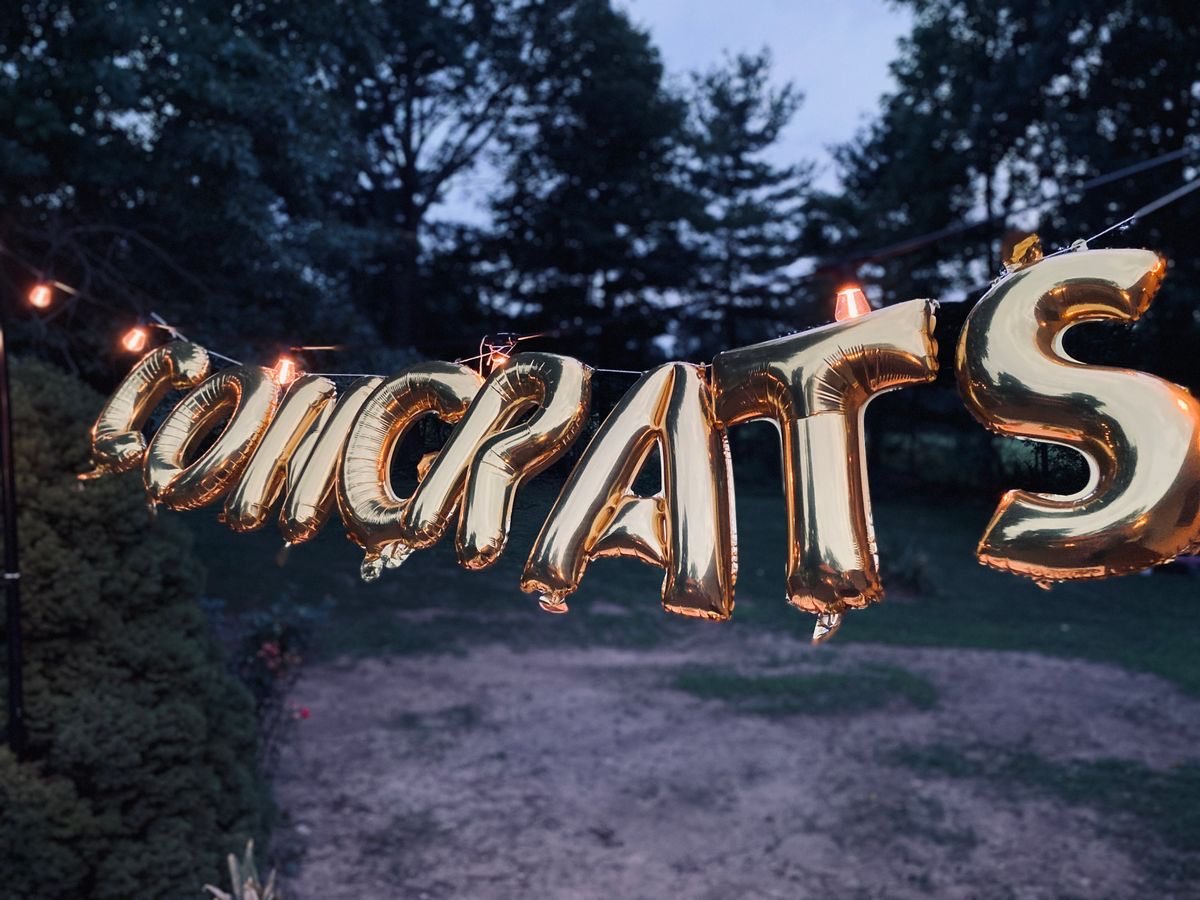 Gold balloon letters spelling Congrats, strung with lights over a backyard at dusk