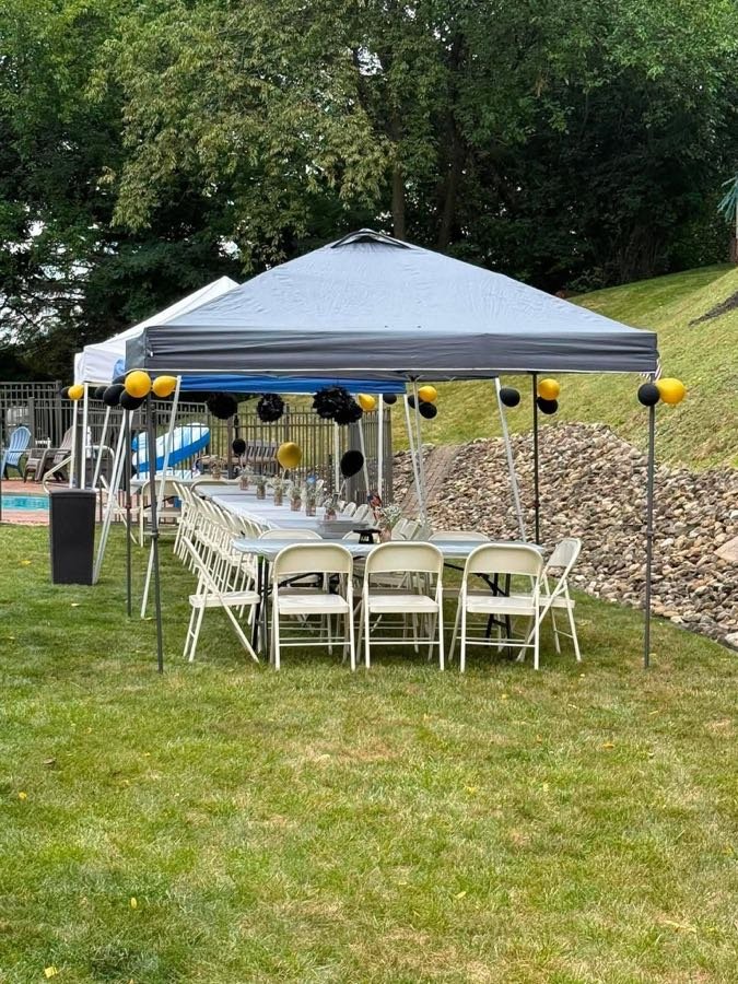 Rows of folding chairs at long tables under a grey canopy with black and gold balloons, set up for a backyard birthday party