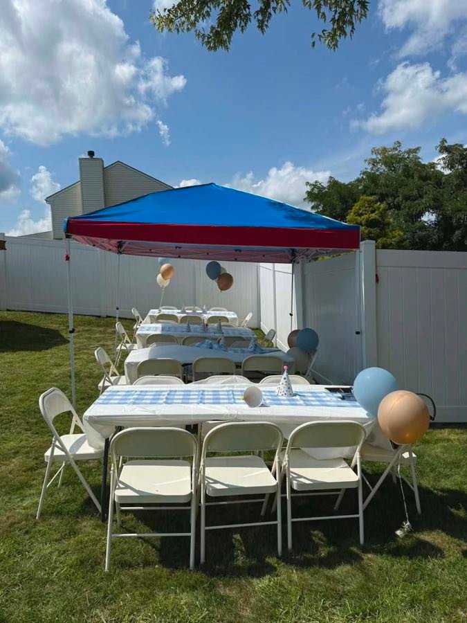 Folding tables with blue gingham runners, party hats and balloons, and folding chairs on a lawn under a canopy, set up for a backyard birthday party