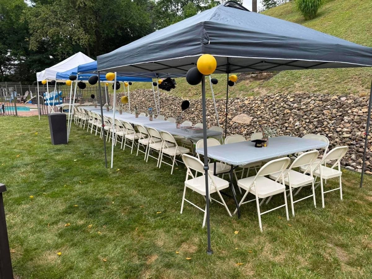 A long run of folding tables with folding chairs along both sides under pop-up canopies, set up for a backyard birthday party