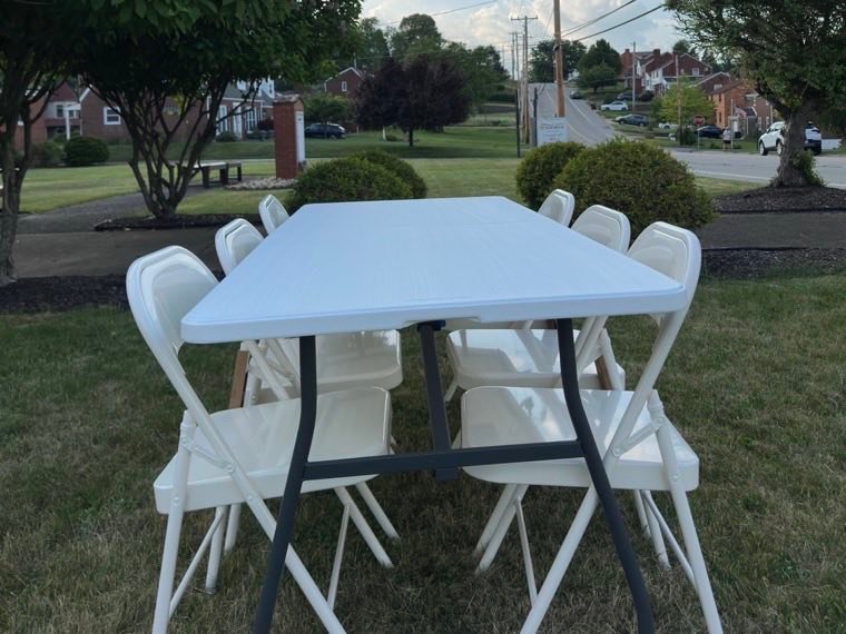 A white six foot rectangular folding table with white folding chairs around it on a lawn